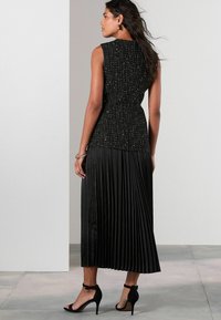 Czarny top bez rękawów z teksturowanej tkaniny, zestawiony z plisowaną czarną spódnicą maxi. Strój ma dopasowany krój i delikatny połysk. Całość uzupełniają czarne szpilki.