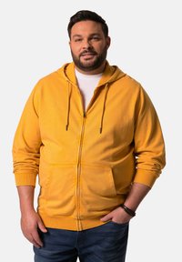Sudadera con cremallera de color amarillo brillante, con un bolsillo frontal, capucha con cordón y puños acanalados, combinada con jeans azules. Tejido suave y texturizado.