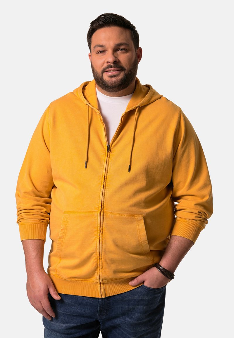 Sudadera con cremallera de color amarillo brillante, con un bolsillo frontal, capucha con cordón y puños acanalados, combinada con jeans azules. Tejido suave y texturizado.
