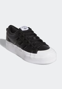 Černá kožená teniska s texturovaným svrškem, bílou gumovou platformou a černými tkaničkami. Obsahuje perforované boční pruhy a štítek s logem.