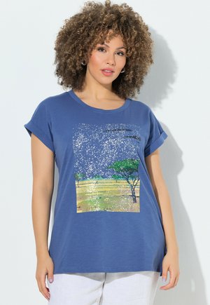 Mujer con cabello rizado que lleva una camiseta azul con un estampado de paisaje natural y pantalones blancos sobre un fondo claro y liso.