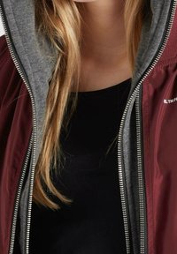 Vínová bunda se šedou kapucí a vícevrstvým designem, vybavená zipem a hladkým vnějším povrchem. Černý top pod ní.