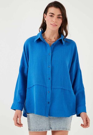 Blue button-up shirt with a textured fabric and long sleeves. Features a slightly oversized fit and a hem that curves at the sides.