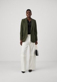 Blazer su misura verde oliva, top aderente nero con dettaglio a cuore, pantaloni bianchi a gamba larga con tasche cargo, tacchi a punta neri e borsa piccola.