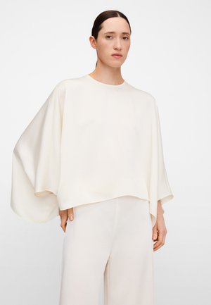 Femme aux cheveux foncés portant un haut blanc ample et fluide à manches larges et un pantalon blanc assorti, debout sur fond uni.