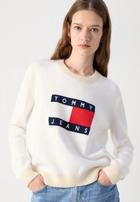 Hvit strikket genser med rund hals, med et logo-design av "TOMMY JEANS" i marineblått og rødt. Ribbestrikkede mansjetter og hem i kremfarge.
