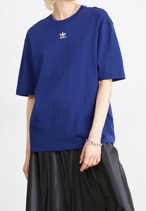Osoba ubrana w niebieską koszulkę Adidas z małym białym logo, czarną spódnicę, srebrną bransoletkę z łańcuszkiem i naszyjnik na jednolitym białym tle.