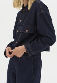 Henkilö, joka käyttää tummansinisestä denimmistä valmistettua takkia ja housuja, joissa on kontrastivärisiä saumoja, säätää housujen vyötäröaluetta yksinkertaista taustaa vasten.