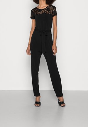 Vrouw gekleed in een zwart jumpsuit met korte kanten mouwen en een borstpaneel, een ceintuur met strik in de taille en zwarte open-toe hoge hakken.