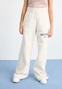 Bílé, široké tepláky s vysokým pasem a šňůrkou. Na levém stehně je vyšitý logo "NIKE" v tmavě modré barvě. Měkká textura.