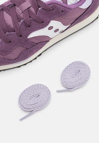 Chaussure de sport violette en daim et mesh, arborant un logo blanc, des lacets plats et deux lacets lilas roulés à côté.