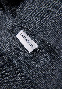 Grijs textiel met een gestructureerde weving, met een wit label met de tekst "LINDBERGH" in de naad genaaid. Close-up benadrukt het detail van het materiaal.