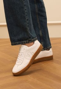 White leather sneakers with a textured upper, featuring white laces, a rubber gum sole, and subtle branding detail on the side.