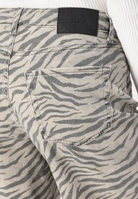 Pantalones de mezclilla beige con un patrón de rayas de tigre negro, con una etiqueta negra en la cinturilla y en los bolsillos traseros. Detalle de superficie texturizada.