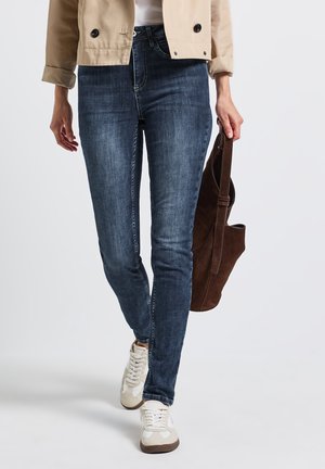 Jean skinny bleu foncé avec une texture délavée, associé à une veste courte beige et des baskets beiges, tout en tenant un sac en daim marron.
