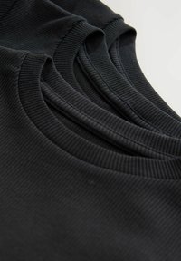 Drei übereinandergestapelte schwarze, gerippte Rundhals-Shirts, die im Nahaufnahme präsentiert werden, wobei die Stoffstruktur und die Kragen Details hervorgehoben werden.
