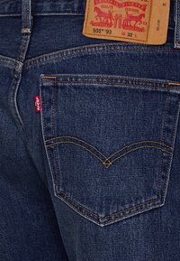 Mörkblå denimjeans med synlig gul sömnad, med en klassisk design för bakfickor och en brun läderetikett med röd text.