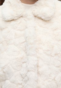 Manteau en fausse fourrure blanche, douce et duveteuse, avec un col arrondi, porté par une personne, montrant la texture et des détails en gros plan.