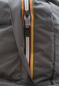 Primo piano di una giacca a vento grigia con cerniera, evidenziata da un bordo in tessuto multicolore a righe e un gancio nero per la cerniera.