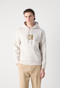 Hanorac cu glugă din amestec de bumbac în bej deschis, având un patch cu logo aurit pe piept și o buzunar kangaroo frontal.