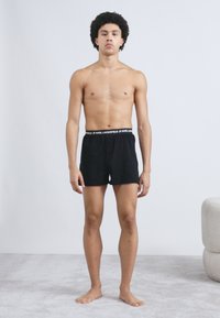 Μαύρα βαμβακερά μποξέρ με ελαστική μέση που φέρει το λογότυπο "KARL LAGERFELD". Το μοντέλο στέκεται με ένα απλό φόντο και υφή στο δάπεδο.