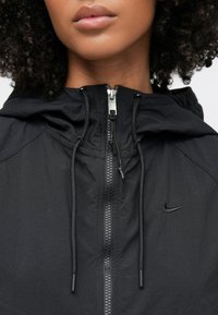 Μαύρο ελαφρύ μπουφάν με κουκούλα, με φερμουάρ μπροστά, διακοσμητικά κορδόνια και μικρό μαύρο λογότυπο Nike στην αριστερή πλευρά.