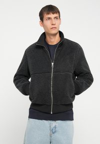 Jachetă bombă neagră din fleece cu guler înalt, închidere cu fermoar, manșete și tiv ribbate. Textură moale cu o siluetă aerodinamică.