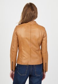 Tan leren jack met een getailleerd ontwerp, voorzien van een opstaande kraag en ritssluitingen bij de manchetten. Gladde textuur en subtiele stikdetails.