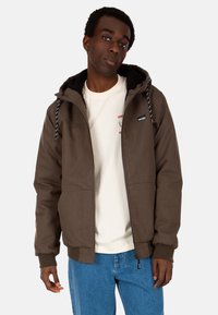 Braune Zip-Jacke mit Kapuze, ausgestattet mit einem Fleece-Innenfutter und Kordelzügen. Getragen über einem cremefarbenen T-Shirt und blauen Jeans.