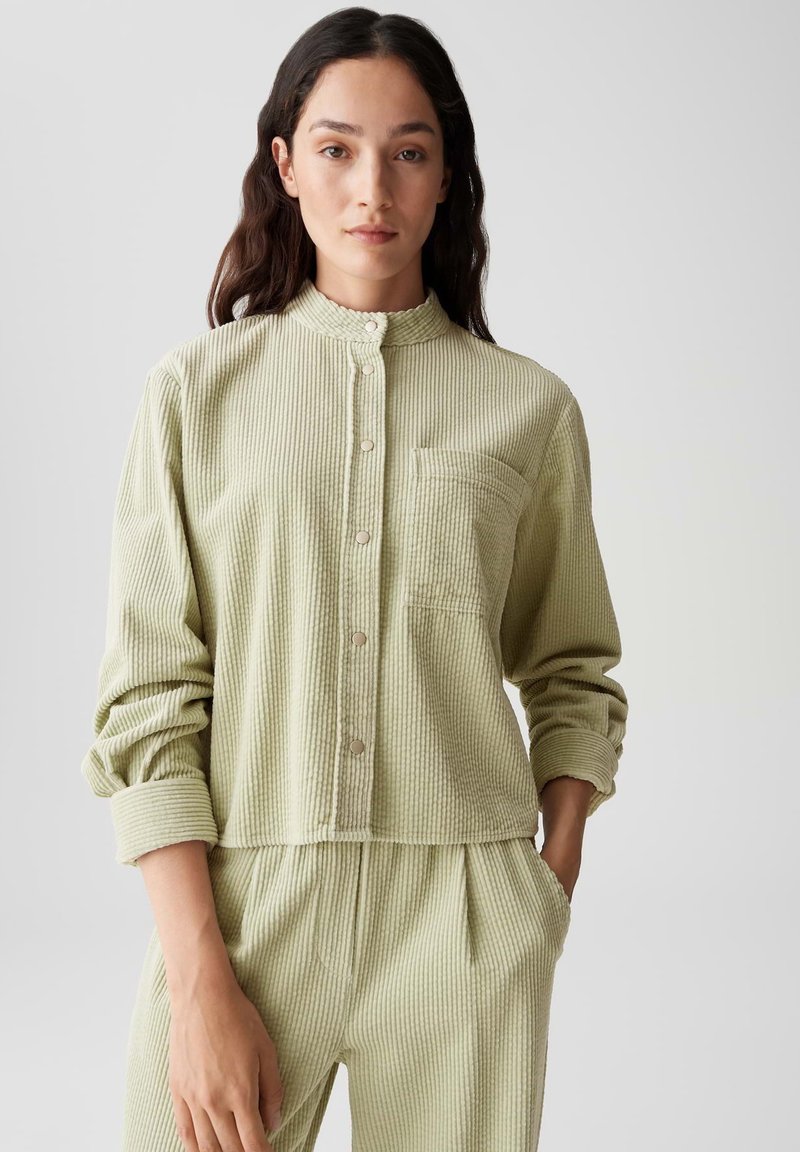 Camicia in corduroy verde chiaro con colletto rotondo, chiusura con bottoni, maniche lunghe e taschino sul petto. Tessuto testurizzato con strisce verticali.