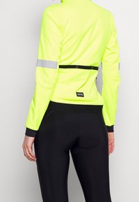 Gul reflexlöpjacka med lång ärm, med ett svart elastiskt midjeband och en logotypapplikation. Kombinerad med svarta åtsittande leggings.