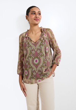 Blusa ligera y estampada en verde con diseños florales en rosa. Presenta un escote en V y mangas tres cuartos. Combinada con pantalones beige.
