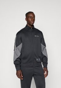 Μαύρο αθλητικό σακάκι με γκρι λεπτομέρειες, κολάρο με φερμουάρ και αντίθετες λευκές ρίγες στα μανίκια. Μαλακή, λεία υφή, με λογότυπο.