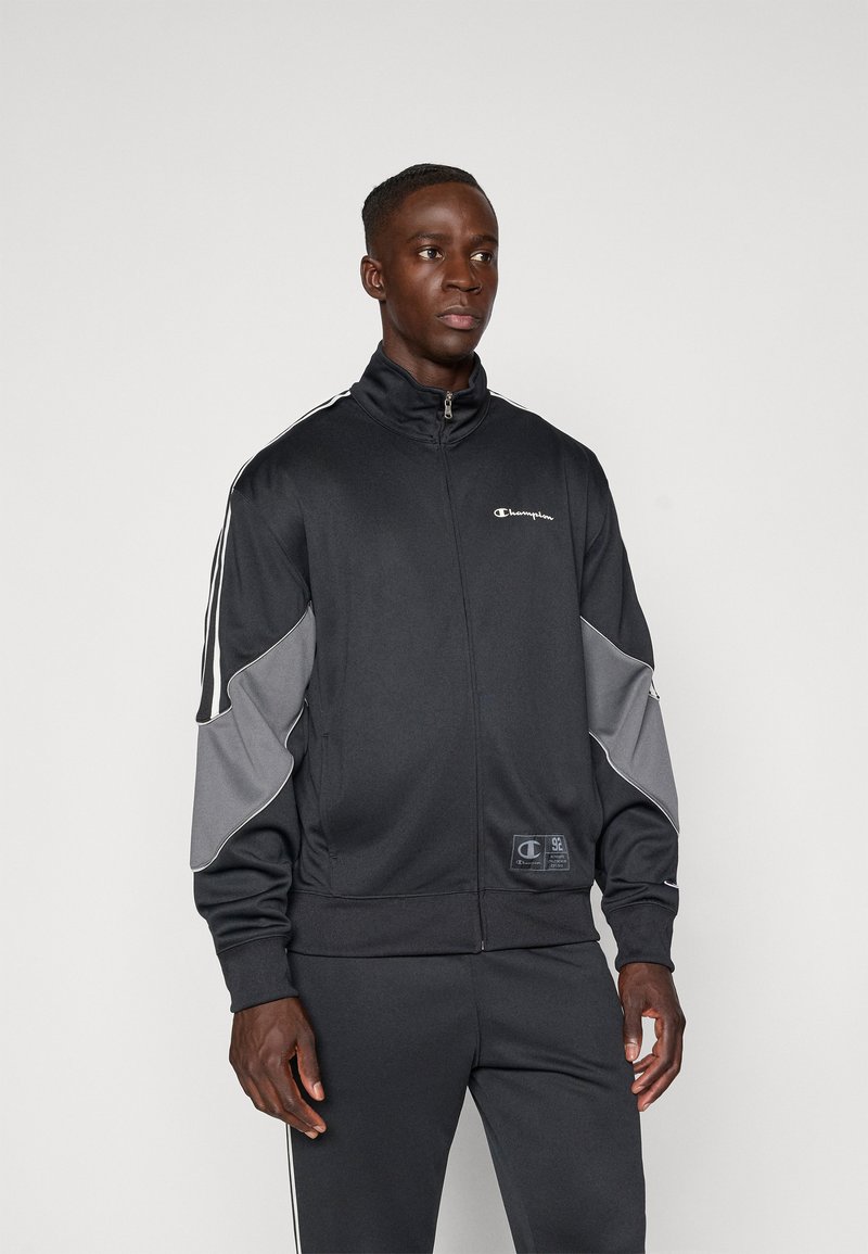 Μαύρο αθλητικό σακάκι με γκρι λεπτομέρειες, κολάρο με φερμουάρ και αντίθετες λευκές ρίγες στα μανίκια. Μαλακή, λεία υφή, με λογότυπο.