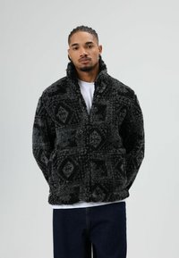 Zachte jas met een zwart-grijs patroon, voorzien van een hoge kraag en twee voorzakken, gedragen over een wit t-shirt en gecombineerd met donkere spijkerbroeken.