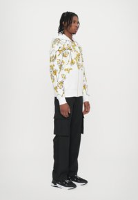 White zip-up hoodie with floral yellow and gray patterns, paired with black cargo pants and black sneakers. Fabric appears soft and lightweight.