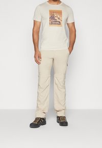 Beige kortærmet T-shirt med grafisk print, sammen med beige zip-off cargobukser og sorte vandresko med orange detaljer.