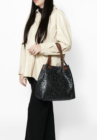 Borsa nera con decorazione floreale in rilievo e manici in pelle marrone, caratterizzata da una forma strutturata e una finitura testurizzata, tenuta da una persona che indossa una camicia chiara.