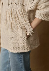 Chlupatý pletený cardigan s otevřeným předním dílem, texturovanými vzory a střapci na lemu. Odpovídající světlým modrým džínsům.