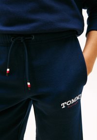 Άτομο που φοράει σκούρο μπλε φόρμα Tommy Hilfiger με κορδόνι, με κόκκινες, λευκές και μαύρες λεπτομέρειες, με το χέρι στην τσέπη.