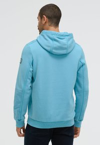 Man wearing light blue hoodie with hood down, facing away, with dark pants against a plain light background.