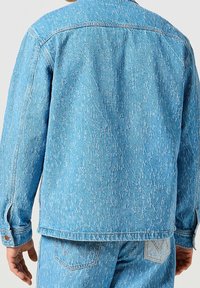 Giacca di jeans azzurro chiaro con maniche lunghe, cuciture visibili e una finitura leggermente strutturata. Presenta un colletto classico e polsini con bottone.