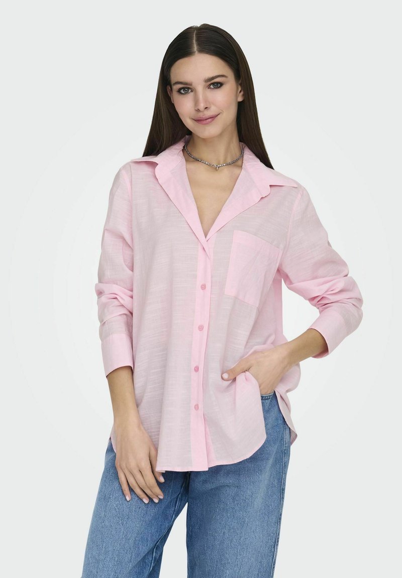 Camisa de botón rosa claro con cuello, mangas largas y un bolsillo en el pecho, hecha de una tela texturizada. Llevada con jeans azules.