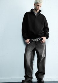 Felpa nera con zip e polsini a coste sopra una camicia a quadri, abbinata a jeans larghi a righe grigie e stivali neri.