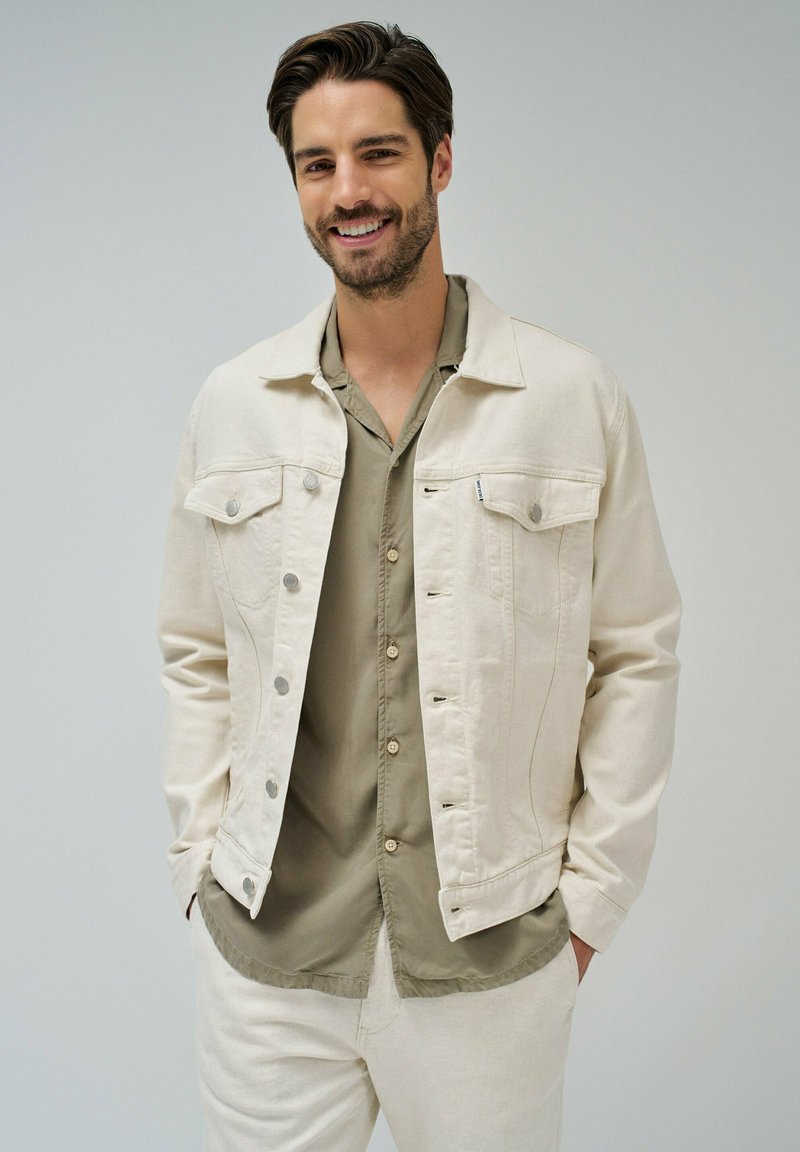 Chaqueta de mezclilla color marfil con dos bolsillos en el pecho, herrajes plateados y un ajuste ligeramente relajado, llevada sobre una camisa de botones verde.