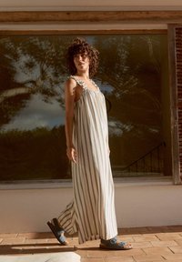 Ärmelloses Maxikleid mit rüschierten Trägern, vertikalen cremefarbenen und grauen Streifen. Getragen mit blauen Sandalen, die über zwei verstellbare Riemen verfügen.