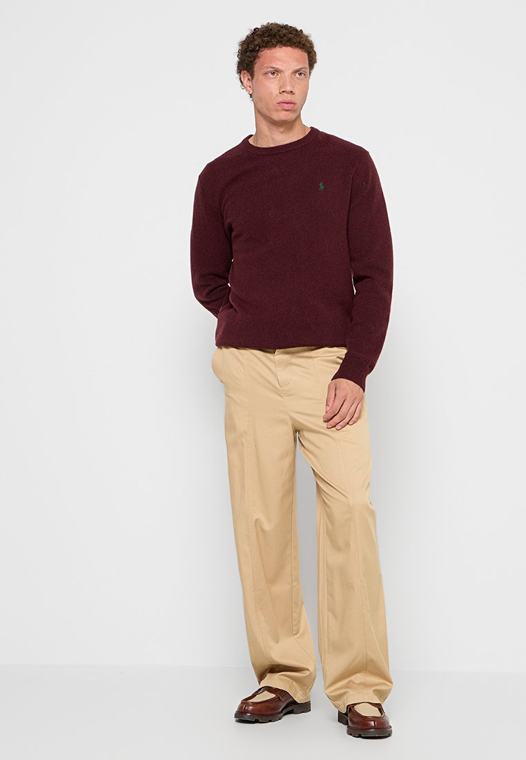 Pull en maille bordeaux à manches longues, associé à un pantalon beige à jambes larges et des chaussures brunes, se tenant devant un fond uni.