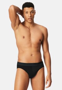 Black mesh men's briefs with a logoed waistband, featuring a snug fit and smooth texture, worn by a model with a neutral expression.