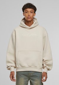 Beige hættetrøje med en frontlomme, ribbede manchetter og hætte med snor. Udstyret med et diskret hvidt logo på brystet. Blød stoftekstur.