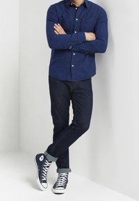 Marinblå rutigt skjorta med knappar, mörk denimjeans och marinblå högskor med vita detaljer. Upprullade ärmar på jeansen.